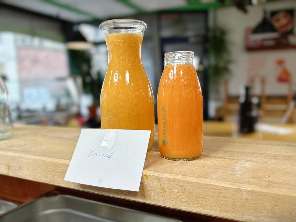 Zwei Glasflaschen mit frisch gepresstem Saft aus dem Kenwood PureJuice Pro stehen auf einer Theke. Ein Zettel mit der Aufschrift „Kenwood“ liegt davor.