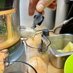 Eine Hand betätigt den Tropfstopp am Saftauslass des Kenwood PureJuice Pro. Saft fließt in einen transparenten Auffangbehälter.