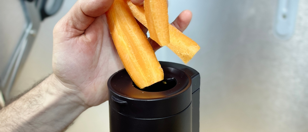 Eine Hand gibt mehrere Karottenstücke in den breiten Einfüllschacht des Panasonic MJ-L700KXE.