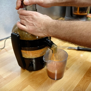 Der Philips HR1832/00 Entsafter im Betrieb presst frischen Karottensaft. Eine Hand drückt das Gemüse durch den Einfüllschacht, während der Saft durch den Auslass in ein Messbecher-Glas fließt.