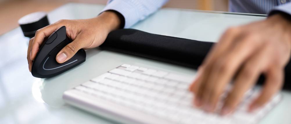 Person sitzt am Schreibtisch, auf dem eine Tastatur liegt, und hält eine ergonomische Maus in der Hand.