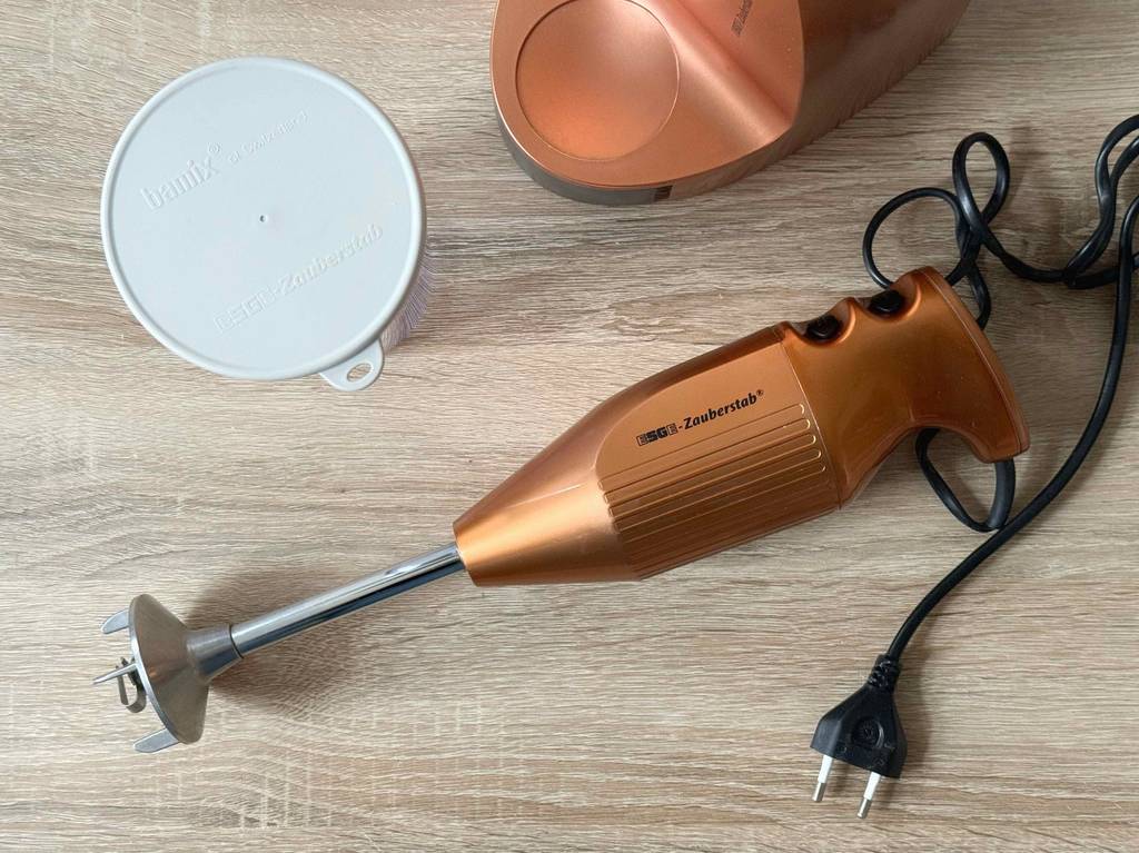 Ansicht von oben auf einen getesteten Esge-Zauberstab im Kupfer-Design mit Handgriff inklusive schwarzer Geschwindigkeitsregler, einem schwarzen Kabel, des Ständers und eines weißen Deckels auf einer Holzfläche.