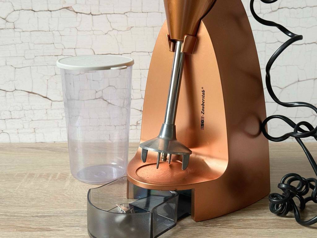 Ein getesteter Esge Zauberstab im bronzefarbenen Design inklusive Zubehörbehälter im Standfuß sowie ein verschließbarer Messbecher stehen auf einer Küchenarbeitsplatte.