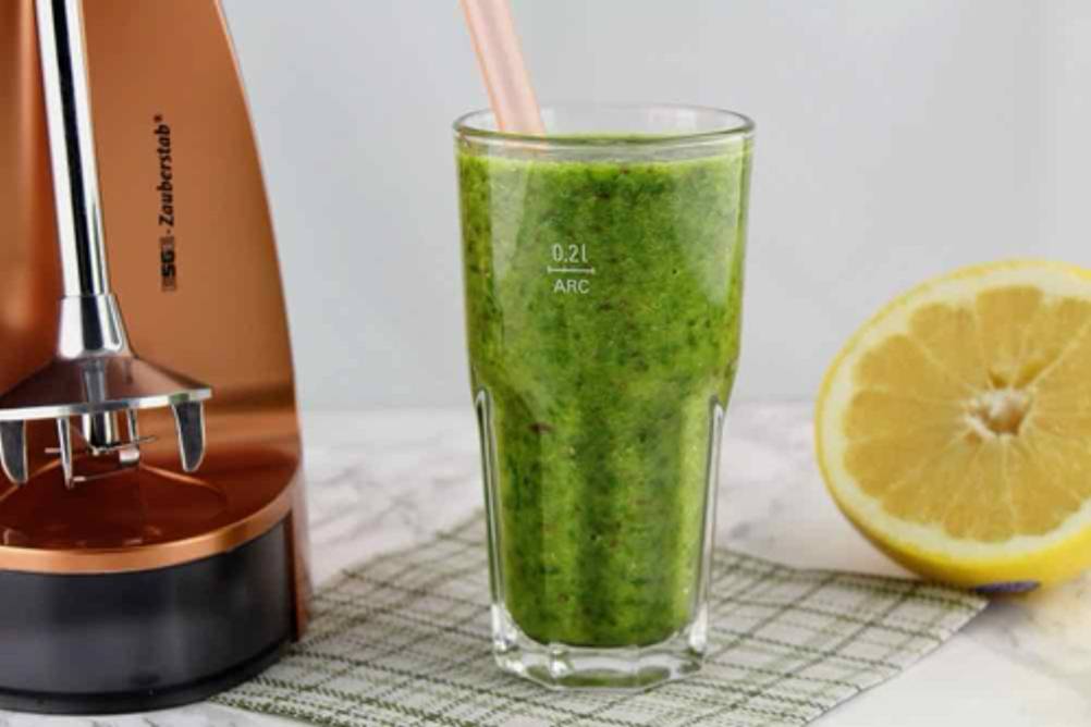 Esge-Zauberstab-Test: Detailansicht eines kupferfarbenen Zauberstab-Ständers inklusive Edelstahl-Rührstab und Klinge neben einem Glas mit grünem Smoothie neben einer haben Zitrone.