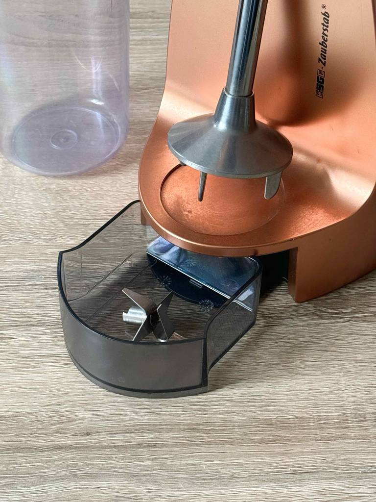Esge-Zauberstab im Test: Detailansicht eines kupferfarbenen Zauberstab-Ständers inklusive einer transparenten Schublade mit Klinge und Edelstahl-Rührstab sowie eines durchsichtigen Kunststoffbehälters.