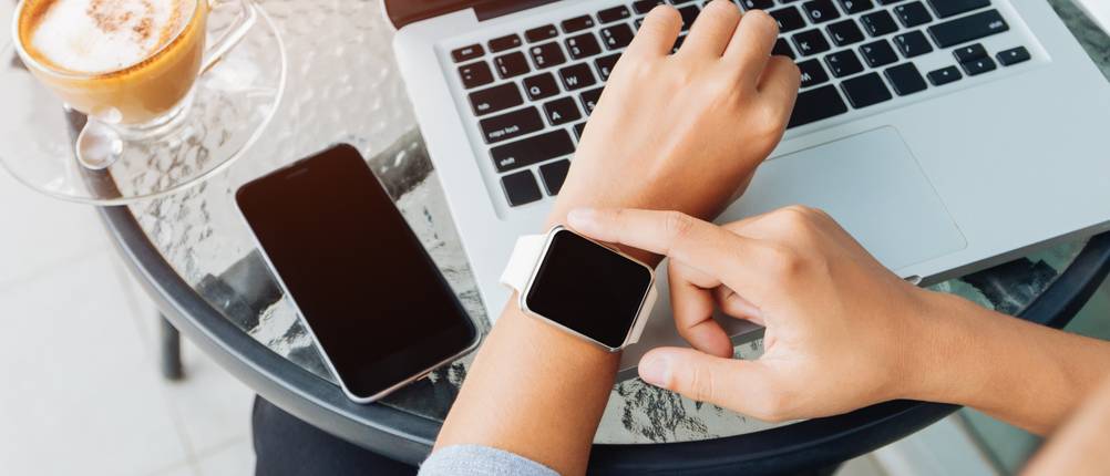 Eine Frau tippt auf ihre Smartwatch. Auf einem Tisch liegen ein Smartphone sowie ein aufgeklappter Laptop.