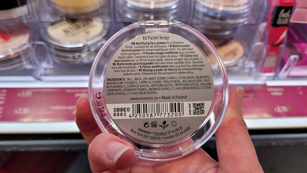 Eine Hand hält eine Dose, mit Sicht auf das hintere Etikett inklusive Produktbeschreibung eines getesteten Essence Puders vor einem Verkaufsregal.