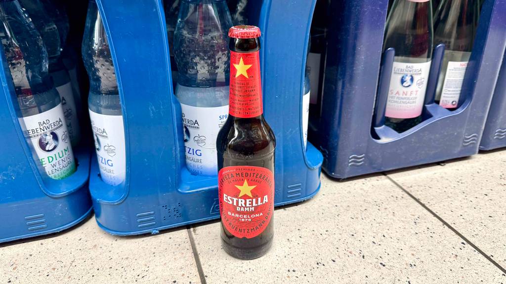 Spanisches Bier getestet: Eine braune Flasche mit rotem Etikett steht vor blauen Getränkekästen.