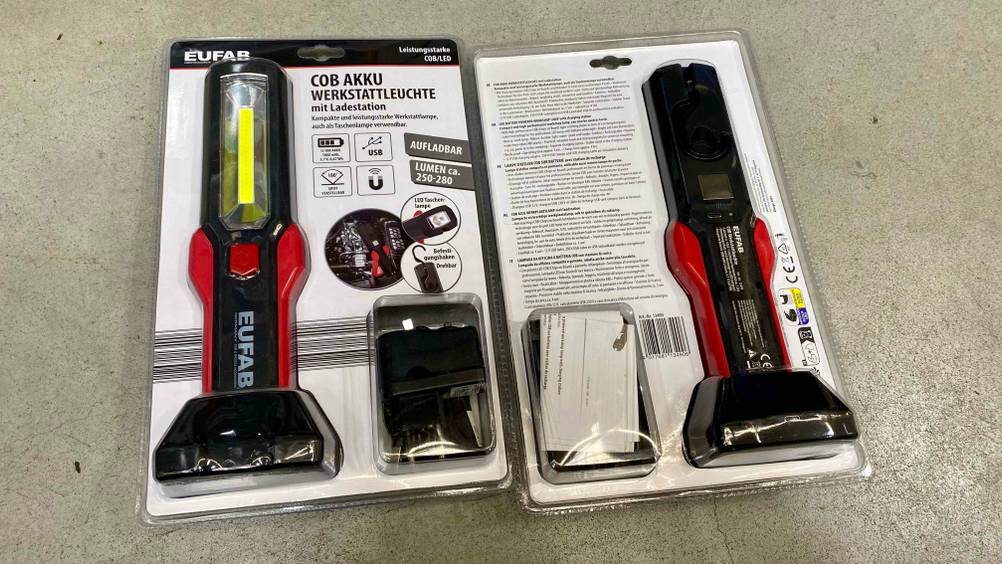 Werkstattlampe getestet: Zwei originalverpackte Geräte auf einem Betonboden, einmal Vorderseite, einmal Rückseite.