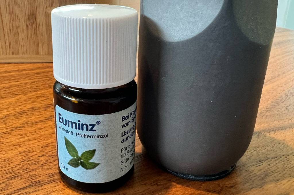 Ein blickdichtes Fläschchen Euminz-Pfefferminzöl im Test steht auf einem Holztisch neben einem Diffusor und einer Grünpflanze im Hintergrund.