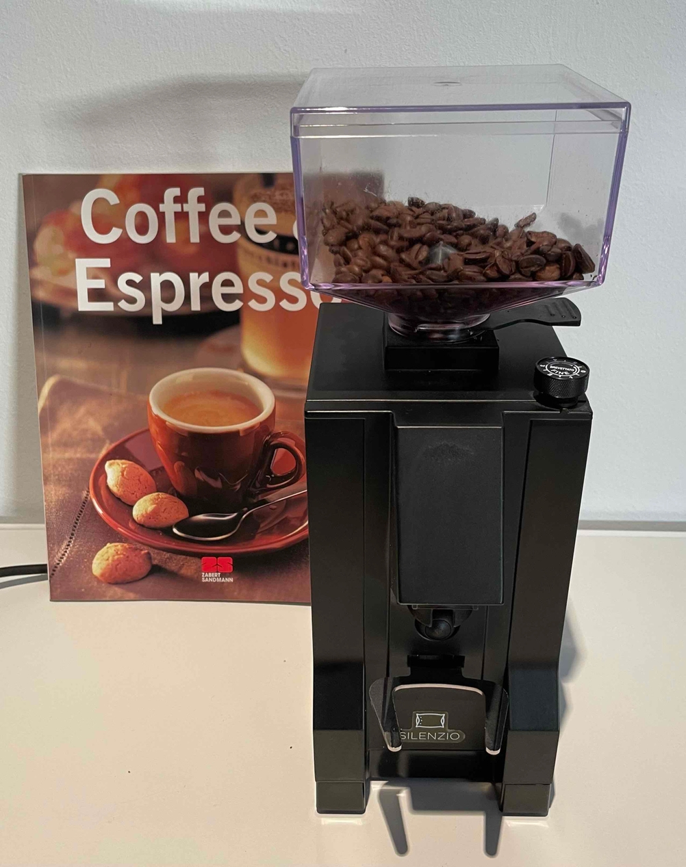 Eine Siebträger-Kaffeemühle im Test steht neben einem Bild mit Coffee-Espresso-Tasse auf einem hellen Tisch.