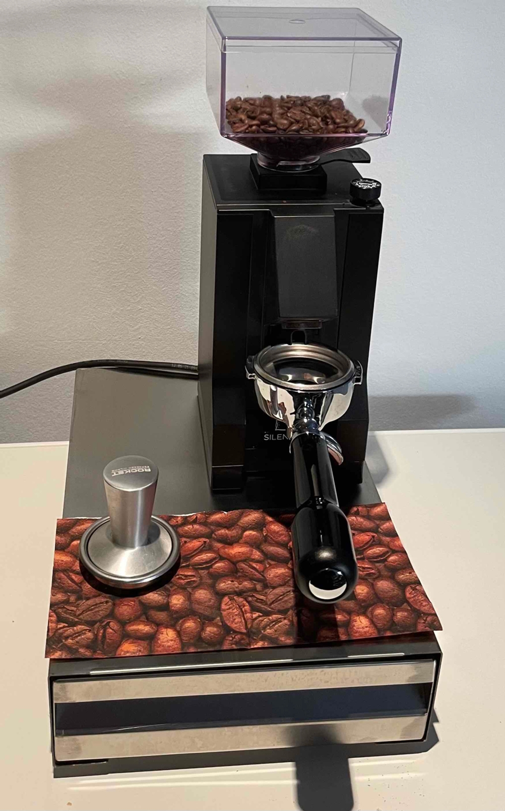 Eine Siebträger-Kaffeemühle steht getestet und inklusive Stopfer auf einer Sudschublade, die sich auf einem hellen Tisch befindet.