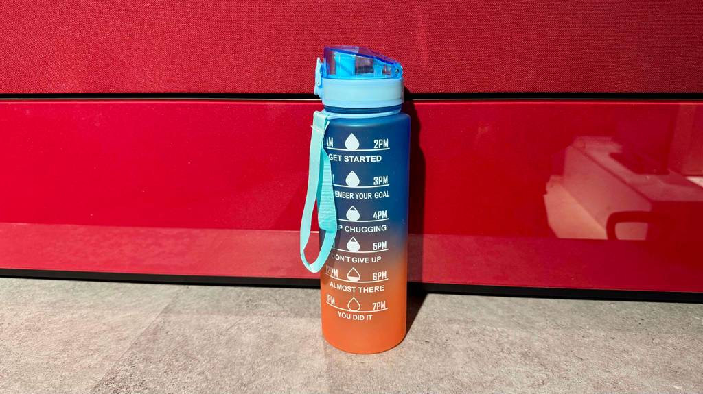 Eine blau-orange Fitness-Trinkflasche im Test steht vor einer roten Wand.