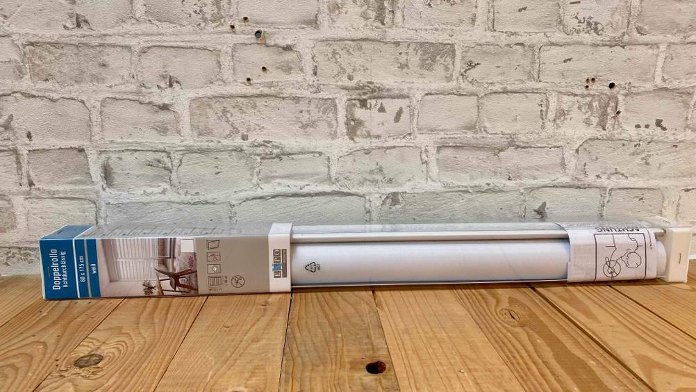 Im Test: Ein verpackter Doppelrollo liegt horizontal auf einer Holzfläche vor einer Steinwand.