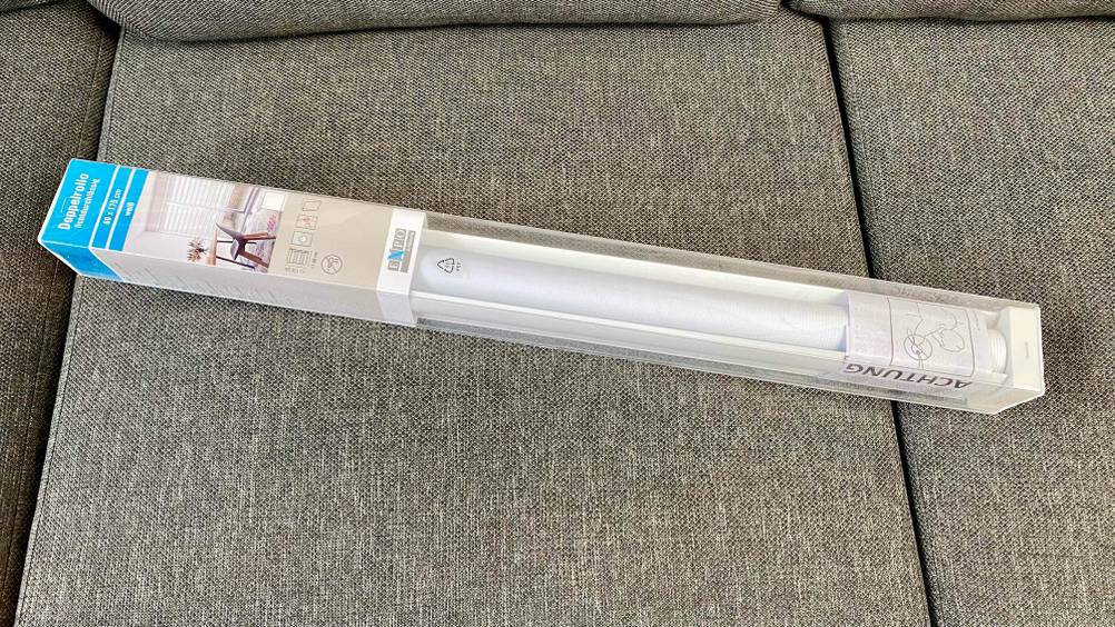 Ein getesteter Doppelrollo in seiner transparenten Verpackung auf einem grauen Polster.