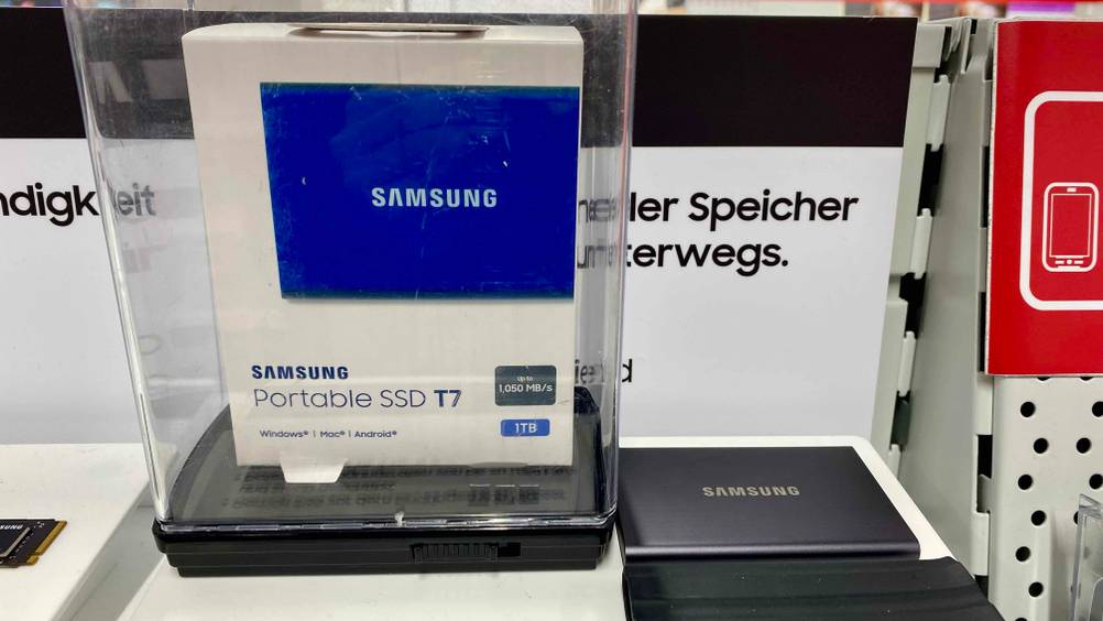 Eine Originalverpackte externe SSD-Festplatte in einem transparenten Gehäuse neben einer auf der Infotafel befestigten.