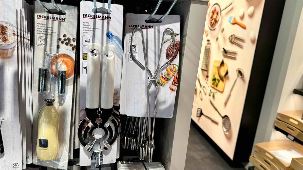 Mehrere getestete Küchenzangen für Fleisch und Gemüse hängen inklusive Produktpappe an einem Verkaufsstand neben weiteren Küchenutensilien.