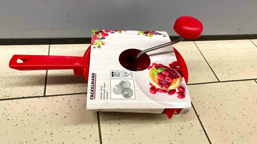 Eine rote getestete Passiermühle von Fackelmann befindet sich in einem weißen Verpackungskarton auf einem beigefarbigen Fliesenboden.