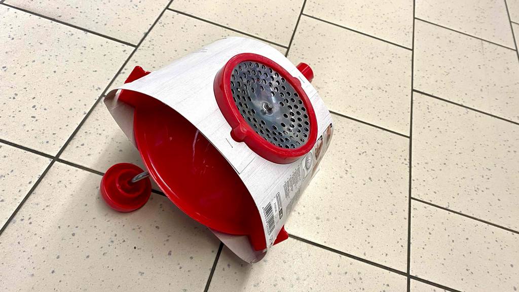 Eine rote getestete Passiermühle liegt, mit Fokus auf die Metall-Lochscheibe und die integrierte Kurbel, in einem weißen Verpackungskarton auf beigefarbenen Fliesen.