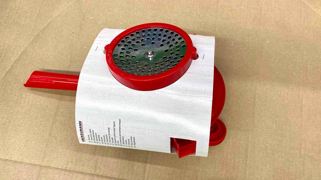 Eine rote Fackelmann-Passiermühle im Test liegt, mit Fokus auf die Metall-Lochscheibe, in ihrem weißen Verpackungskarton auf einer braunen Pappe.