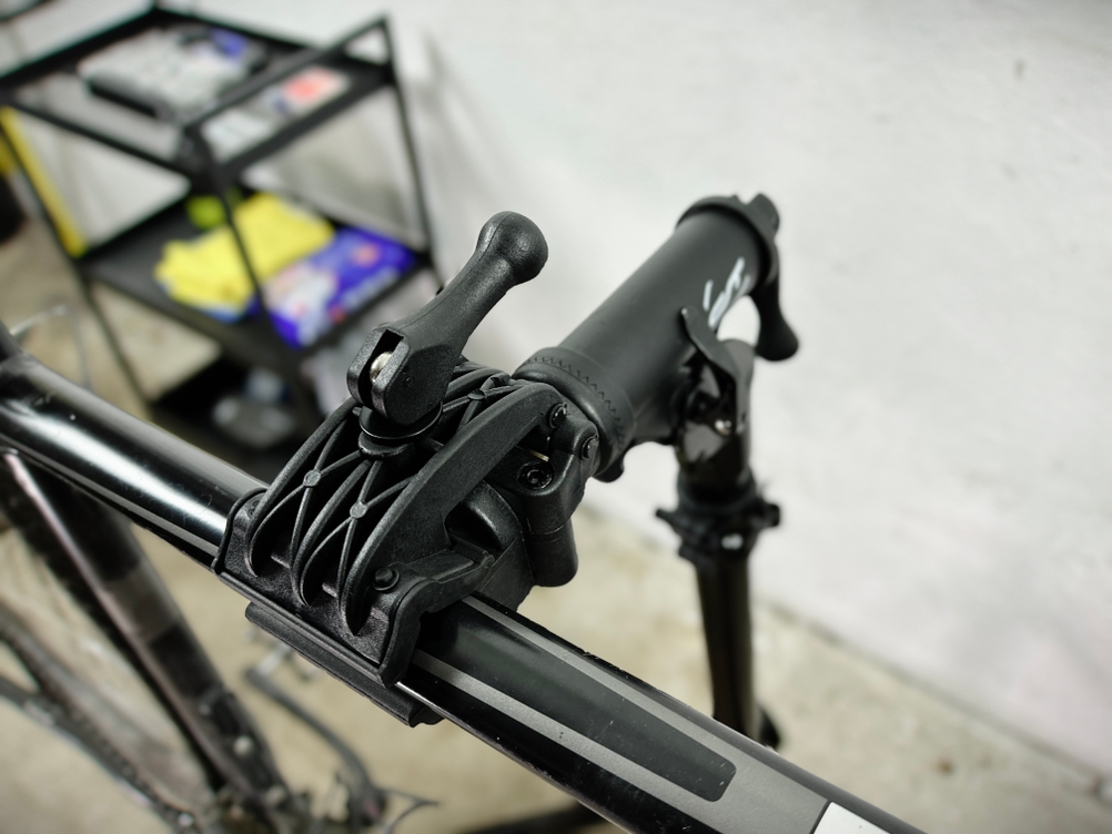 Eine Nahaufnahme des Klemmmechanismus eines Fahrrad-Montageständers, der einen Fahrradrahmen fixiert. Im Hintergrund ist eine Werkstattumgebung mit Regalen zu sehen.