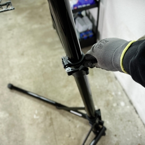 Eine Person verstellt die Arbeitshöhe des Montageständers mit einem Schnellspanner. Der Mechanismus ermöglicht eine ergonomische Anpassung für Reparaturen.