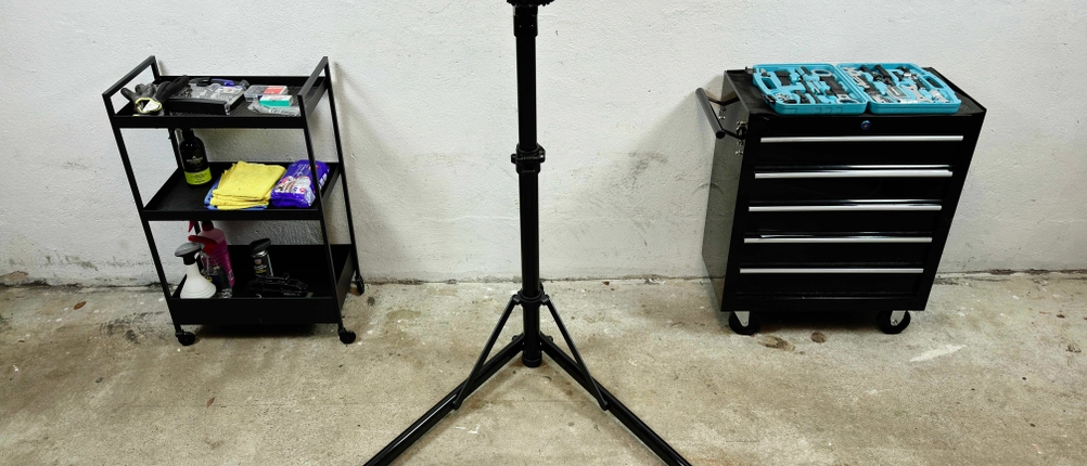 Ein Fahrrad-Montageständer steht aufgebaut in einer Werkstatt. Daneben befinden sich ein Werkzeugwagen und ein Regal mit Pflegeprodukten.