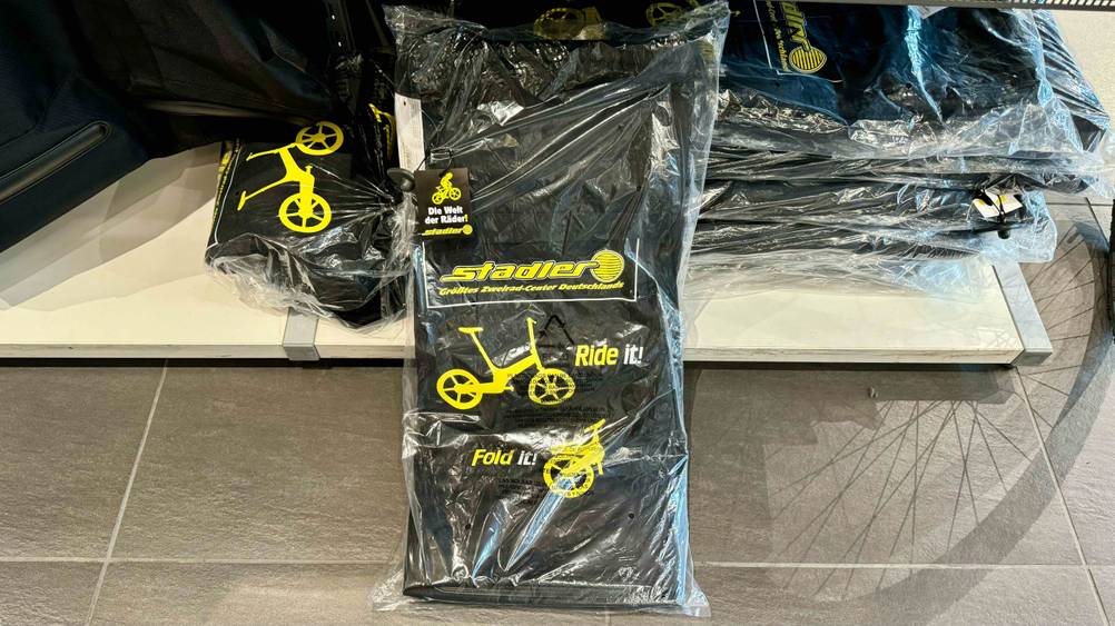 Eine originalverpackte Fahrrad-Transporttasche im Test mit Logo vor einem Regal liegend.