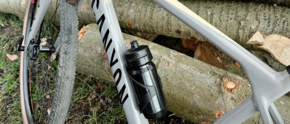 Eine schwarze BBB Cycling Wasserflasche sitzt sicher in der Halterung eines Canyon-Fahrrads. Im Hintergrund sind gestapelte Baumstämme zu sehen.
