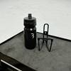 Die BBB Cycling Wasserflasche und der passende Flaschenhalter liegen nebeneinander auf einer dunklen Werkbank. Der Halter ist aus Metall und hat eine schlichte Form.