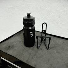Die BBB Cycling Wasserflasche und der passende Flaschenhalter liegen nebeneinander auf einer dunklen Werkbank. Der Halter ist aus Metall und hat eine schlichte Form.