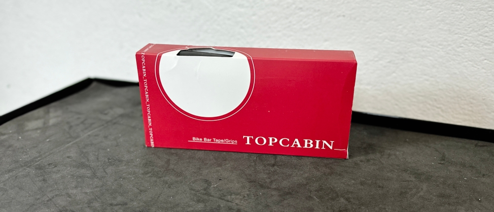 Die Verpackung der Topcabin-Fahrradgriffe steht aufrecht auf einer Werkbank. Der Schriftzug ist klar erkennbar.