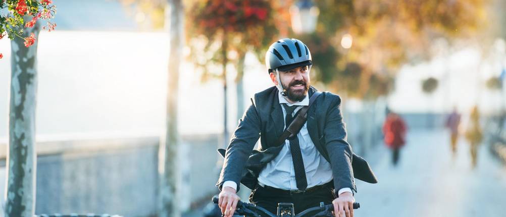 Fahrradhelm-Herren-Test: Businessmann mit Anzug und Krawatte radelt mit blauem Helm eine Baumallee in städtischem Umfeld entlang