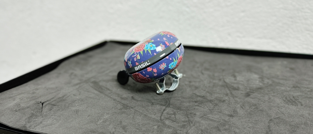 Die Basil-Big-Bell-Bloom-Fahrradklingel mit floralem Design liegt auf einer dunklen Oberfläche. Die Klingel hat ein großes, rundes Gehäuse mit farbenfrohem Muster.