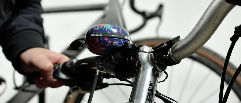 Die Basil Big Bell Bloom wird mit einem Schraubendreher am Fahrradlenker befestigt. Zwei Hände ziehen die Schraube an, um die Klingel zu fixieren.