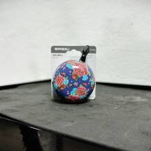 Die Basil Big Bell Bloom Fahrradklingel in ihrer Originalverpackung steht auf einer dunklen Oberfläche. Die Klingel hat ein auffälliges Blumenmuster auf blauem Hintergrund.