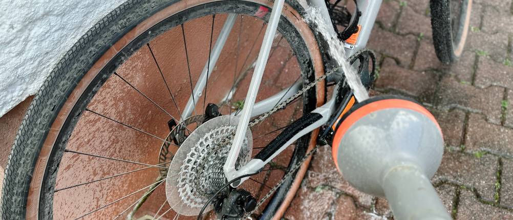 Ein verschmutztes Ritzelpaket wird mit Wasser abgespritzt. Das Fahrrad steht auf einem Pflasterboden vor einer weißen Wand.