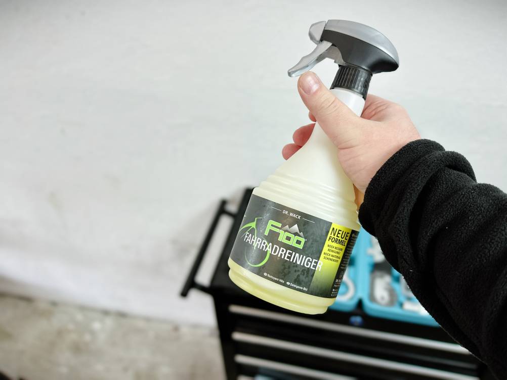 Die Flasche des Dr.-Wack-F100-Fahrradreinigers wird mit der rechten Hand gehalten. Im Hintergrund steht ein geöffneter Werkstattwagen mit Werkzeugeinsatz.