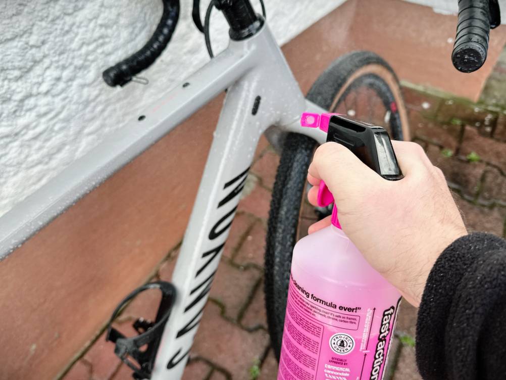 Ein Fahrradrahmen wird im Außenbereich mit dem Muc-Off-Fahrradreiniger eingesprüht. Der Reiniger trifft auf das Oberrohr eines grauen Gravelbikes.