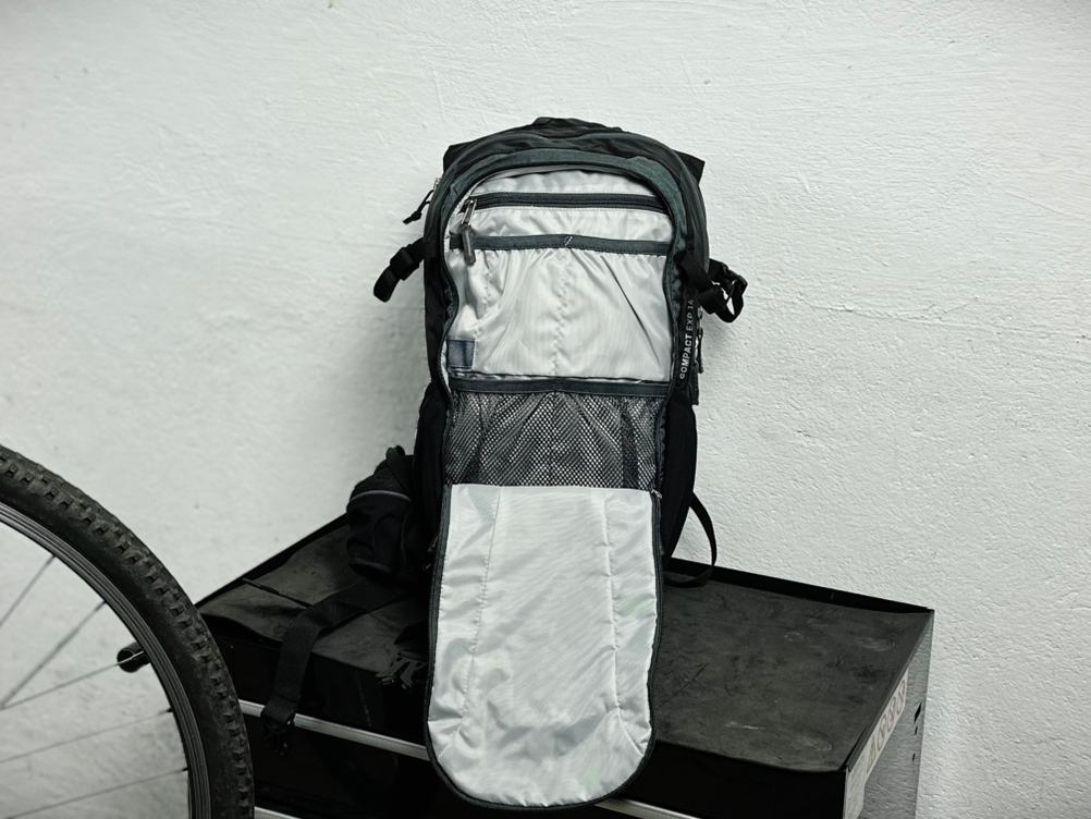 Die Vorderseite des Deuter Compact EXP 14 ist geöffnet und zeigt mehrere Organizer-Fächer im Innenbereich. Die Tasche lehnt an einer weißen Wand.