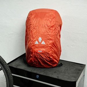 Der Vaude Tremalzo 22 ist mit einer roten Regenhülle bedeckt. Das Vaude-Logo ist auf der Hülle zu sehen.