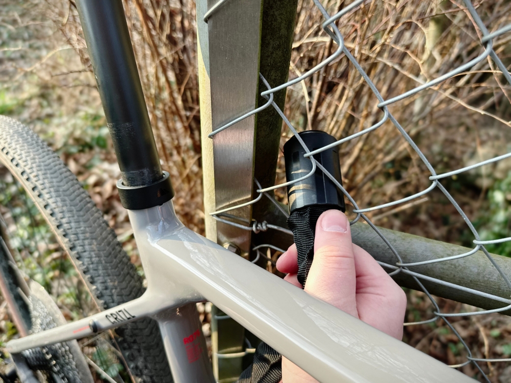 Eine Person führt das Abus CityChain 1010 durch einen Metallzaun. Das Schloss wird um den Rahmen und einen Zaunpfosten gelegt.