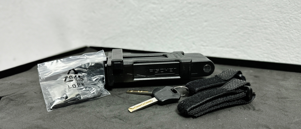 Der Lieferumfang des Fischer-Faltschlosses umfasst zwei Schlüssel, eine Halterung und Befestigungsmaterial. Die Teile liegen auf einer glatten Oberfläche.