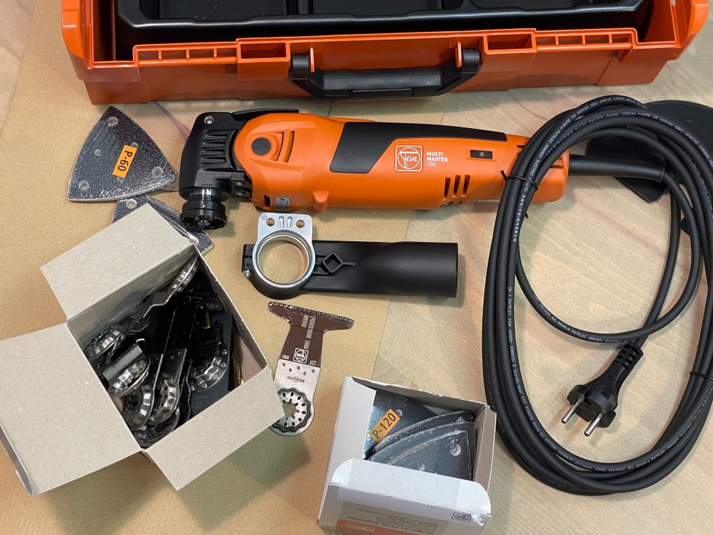 Der Lieferumfang des Fein Multimaster 700 ist vollständig ausgebreitet. Das Gerät liegt neben Koffer, Sägeblättern, Schleifpads und Zubehör.
