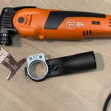 Der Fein Multimaster 700 liegt neben einem Staubsaugeradapter und einem Sägeblatt. Das Kabel ist vollständig aufgerollt.