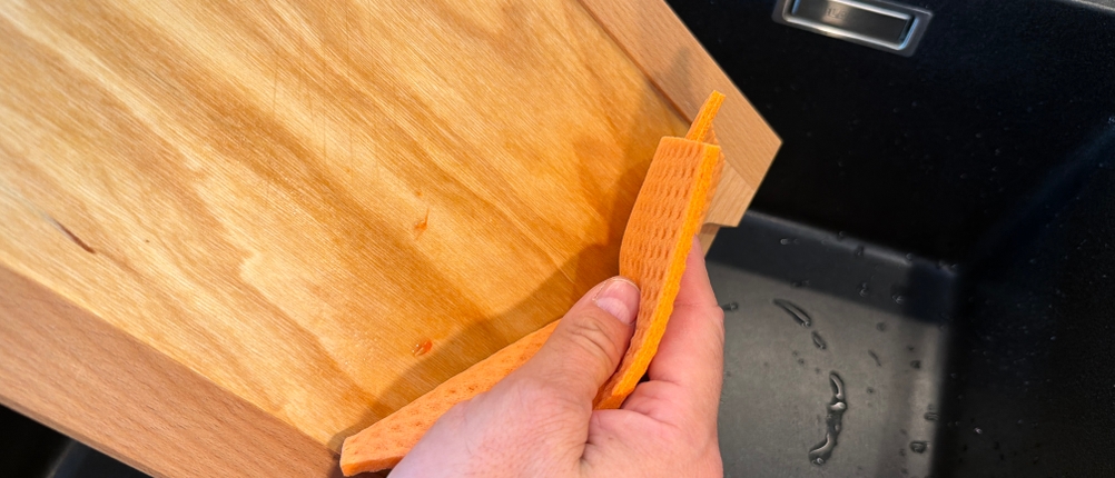 Eine Hand reinigt das Feinholz-Brotschneidebrett mit einem Lappen
