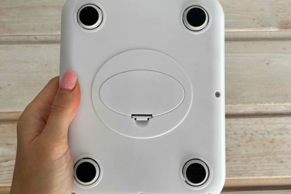 Im Test: Hand hält die Unterseite einer Feinwaage nach vorne vor einer hellen Holzlamellenwand.