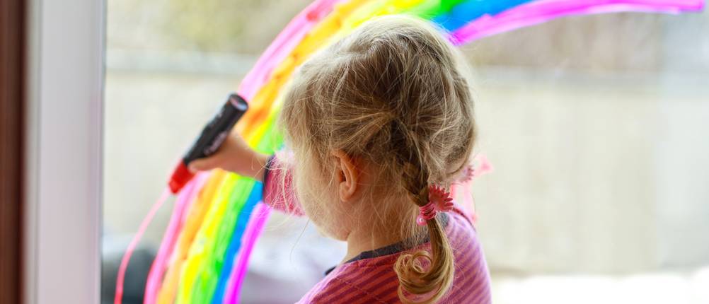 Fenstermalstifte-Test: Kleines Mädchen malt mit Stift einen Regenbogen auf ein Fenster.