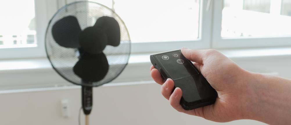 Eine Hand mit Fernbedienung steuert einen Standventilator aus einiger Entfernung.