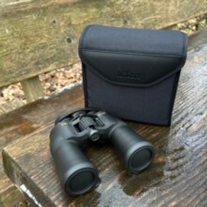 Das Nikon-Aculon-A211-12x50-Fernglas liegt neben seiner schwarzen Tragetasche auf einer Holzbank. Beide sind für den Outdoor-Einsatz konzipiert.